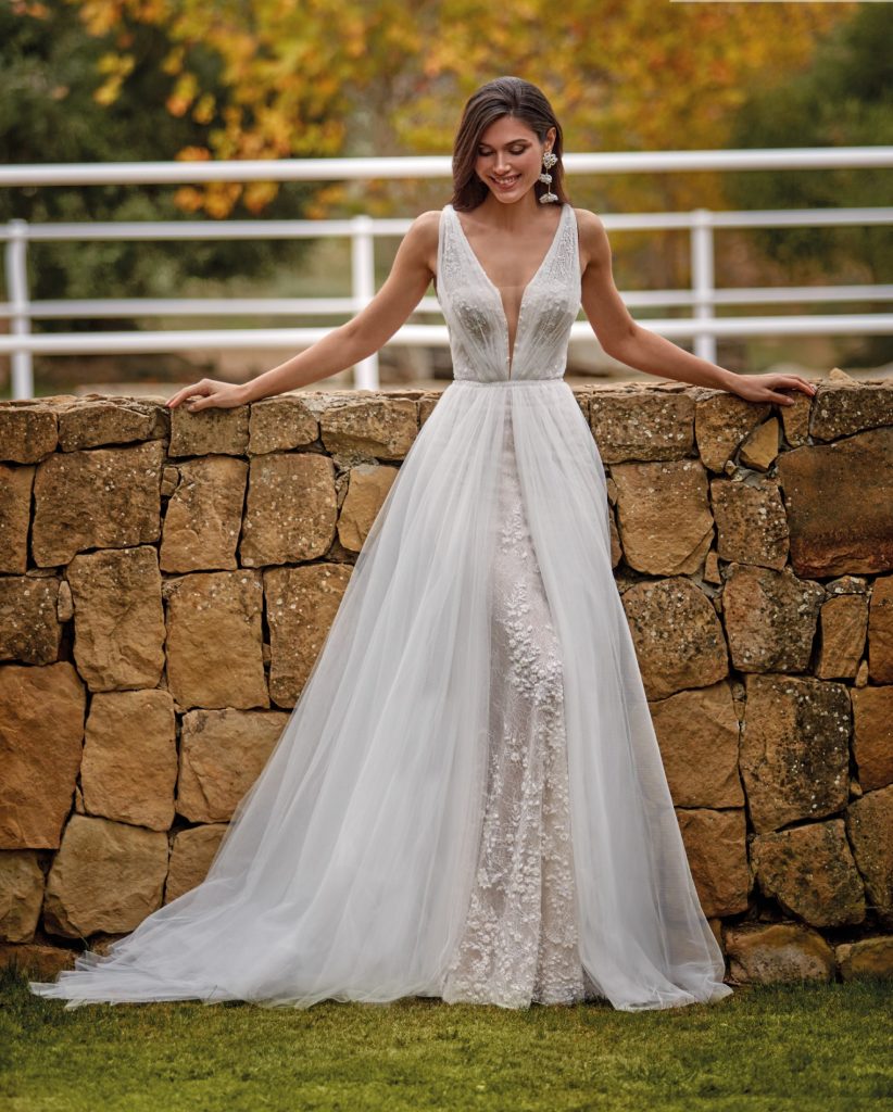 In questa foto si vede un abito da sposa firmato Valerio Luna che rappresenta una delle tendenze sposa 2026 , con profondo scollo a V e gonna in tulle sovrapposta a un raffinato ricamo floreale realizzato in pizzo. Il vestito ha una linea dritta che slancia la figura, mentre lo strato di tulle aggiunge volume e leggerezza. L’effetto finale è etereo e romantico, perfetto per un matrimonio all’aperto.