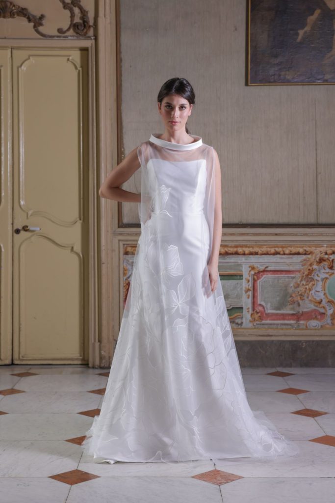 In questa foto si vede un abito da sposa elegante e minimale firmato Atelier Benedetta che rappresenta una delle tendenze sposa 2026 . Il vestito è realizzato in tessuto bianco liscio con sovrapposizione in organza trasparente decorata da delicati motivi floreali in rilievo. Il design è dritto e leggero, con collo morbido a scialle e spalle scoperte, per un effetto raffinato e contemporaneo. L’abito è perfetto per una sposa dal gusto sofisticato.