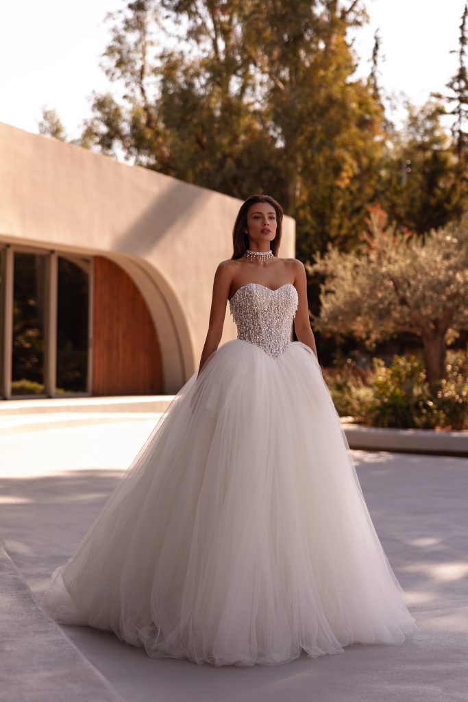 In questa foto si vede un abito da sposa principesco che rappresenta una delle tendenze sposa 2026  firmato Demetrios con corpetto a cuore tempestato di perline e cristalli, che scendono fino al punto vita a punta. La gonna è ampia e vaporosa in tulle bianco latte, creando un perfetto equilibrio tra brillantezza e leggerezza. Il look è completato da una collana-girocollo in pendant con il bustino.