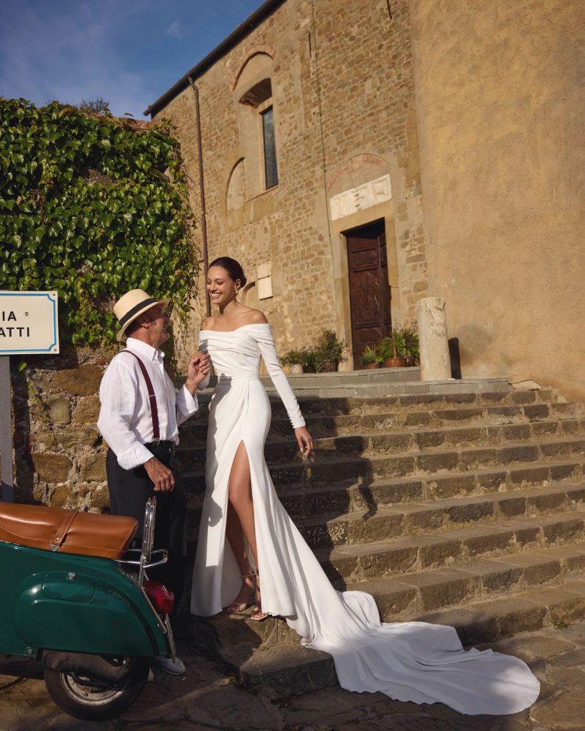 In questa foto, la sposa indossa un abito a sirena bianco con maniche lunghe e scollo a barca drappeggiato che che rappresenta una delle tendenze sposa 2026 e lascia scoperte le spalle. L’abito, aderente fino al ginocchio, si apre in una gonna fluida con uno spacco laterale sulla gamba e termina con un lungo strascico. Il tessuto scivolato e il taglio pulito esaltano la silhouette con eleganza contemporanea.