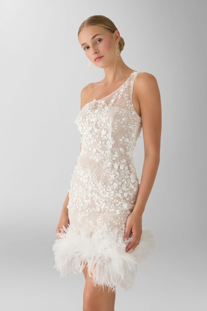 In questa foto, una modella indossa un abito da sposa corto monospalla interamente ricamato con delicati motivi floreali in rilievo. Il vestito, aderente e raffinato, è impreziosito da un bordo di piume bianche vaporose lungo l’orlo, che aggiungono movimento e un tocco di glamour rétro. Lo sfondo neutro esalta la silhouette della modella e i dettagli dell'abito. Questo look rappresenta una delle proposte più originali e femminili tra gli abiti da sposa corti 2026.
