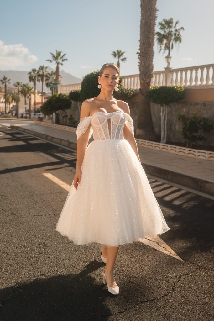 In questa foto, una modella sfila all’aperto sotto il sole indossando un abito da sposa corto, perfetto esempio della tendenza "abiti da sposa corti 2026". Il vestito, lungo fino ai polpacci, presenta un corpino strutturato con stecche a vista e maniche scese, mentre la gonna ampia in tulle scintillante crea un effetto vaporoso e romantico. Il look è completato da scarpe con tacco e orecchini floreali, in un contesto urbano punteggiato da palme e luce naturale.