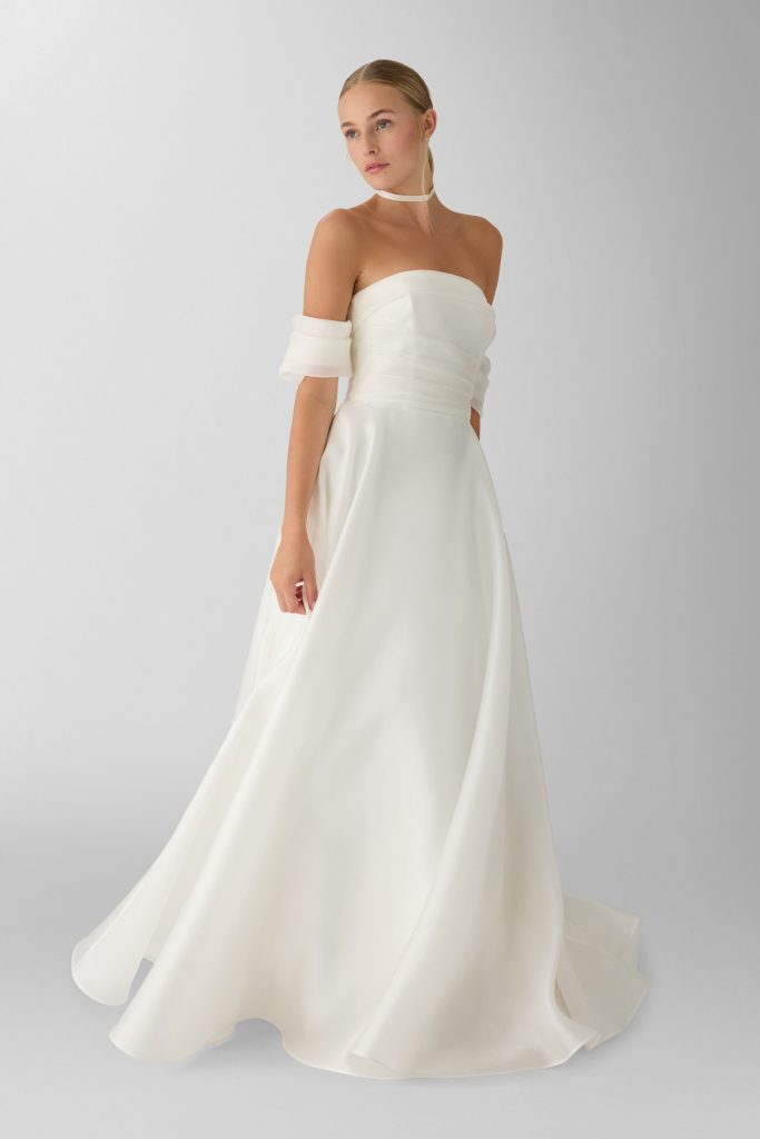 In questa foto, una modella bionda indossa un elegante abito da sposa color avorio con spalle scoperte e maniche morbide che cadono lungo le braccia. Il corpino è semplice e strutturato, con leggere pieghe orizzontali, mentre la gonna scivola con naturalezza verso il pavimento, creando un effetto etereo e fluido. Lo sfondo chiaro valorizza la purezza delle linee e dei tessuti. Questo look rappresenta perfettamente lo stile minimal-chic degli abiti da sposa scivolati 2026, pensati per una sposa moderna e raffinata.