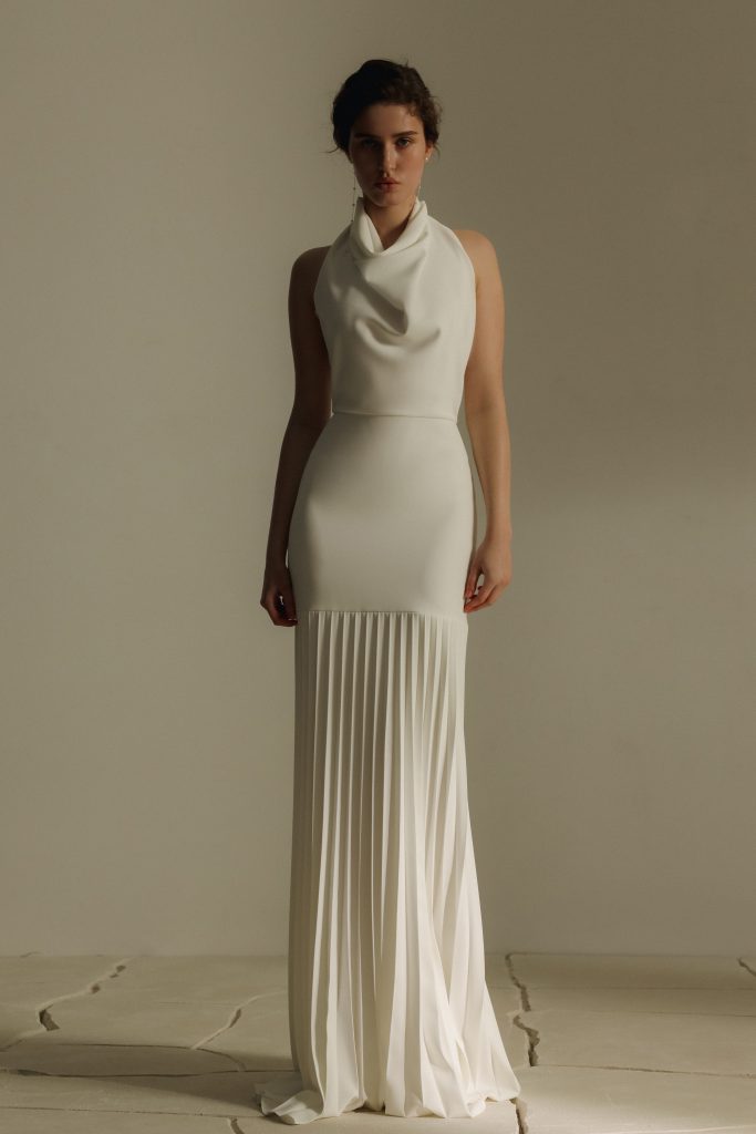In questa foto una modella posa frontalmente indossando un elegante abito bianco da sposa con taglio scivolato. Il vestito presenta un corpetto liscio con scollo morbido drappeggiato e senza maniche, mentre la parte inferiore è caratterizzata da una gonna lunga a pieghe verticali che cade fluida fino a terra. L'atmosfera è minimale e sofisticata, con uno sfondo neutro e pavimento chiaro. L'immagine è parte della selezione “abiti da sposa scivolati 2026”.
