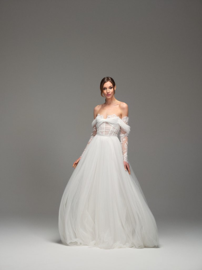 In questa foto, una modella posa in uno studio dal fondale neutro, indossando un abito da sposa dallo stile principesco e raffinato. Il corpetto è trasparente, con maniche lunghe in pizzo e scollo a cuore con drappeggi che scendono sulle braccia. La gonna, ampia ma leggera, scende fluida in tulle vaporoso, rappresentando una delle proposte più scenografiche tra gli abiti da sposa scivolati 2026.