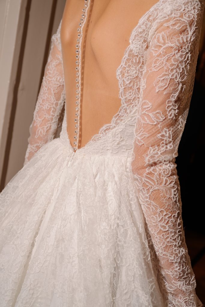 In questa foto, vediamo il dettaglio posteriore di un abito della collezione abiti da sposa Atelier Emé 2026. Il vestito presenta un profondo scollo sulla schiena, bordato da un elegante pizzo floreale. Le maniche lunghe in tulle trasparente sono anch’esse ricamate, e una fila di piccoli bottoni perlati accompagna la schiena fino al punto vita, donando un tocco di raffinatezza classica.