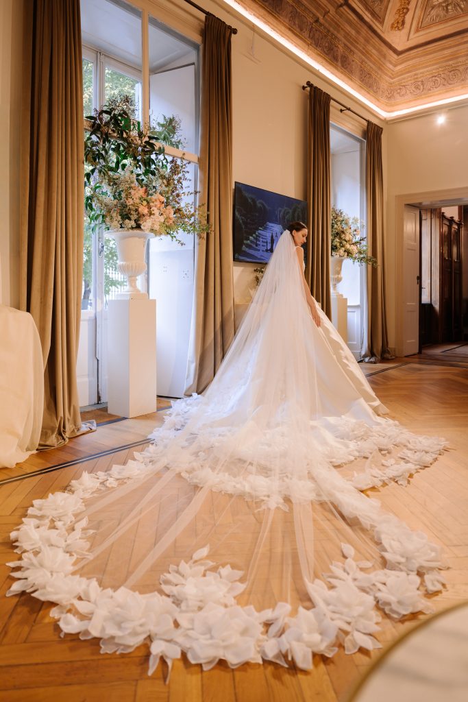 In questa foto, una modella attraversa una sala elegante con soffitto affrescato, indossando un sontuoso abito della collezione abiti da sposa Atelier Emé 2026. Il vestito ha uno strascico lunghissimo in tulle, decorato da una cornice di grandi fiori tridimensionali che sembrano sbocciare sul pavimento, creando un effetto scenografico e fiabesco.