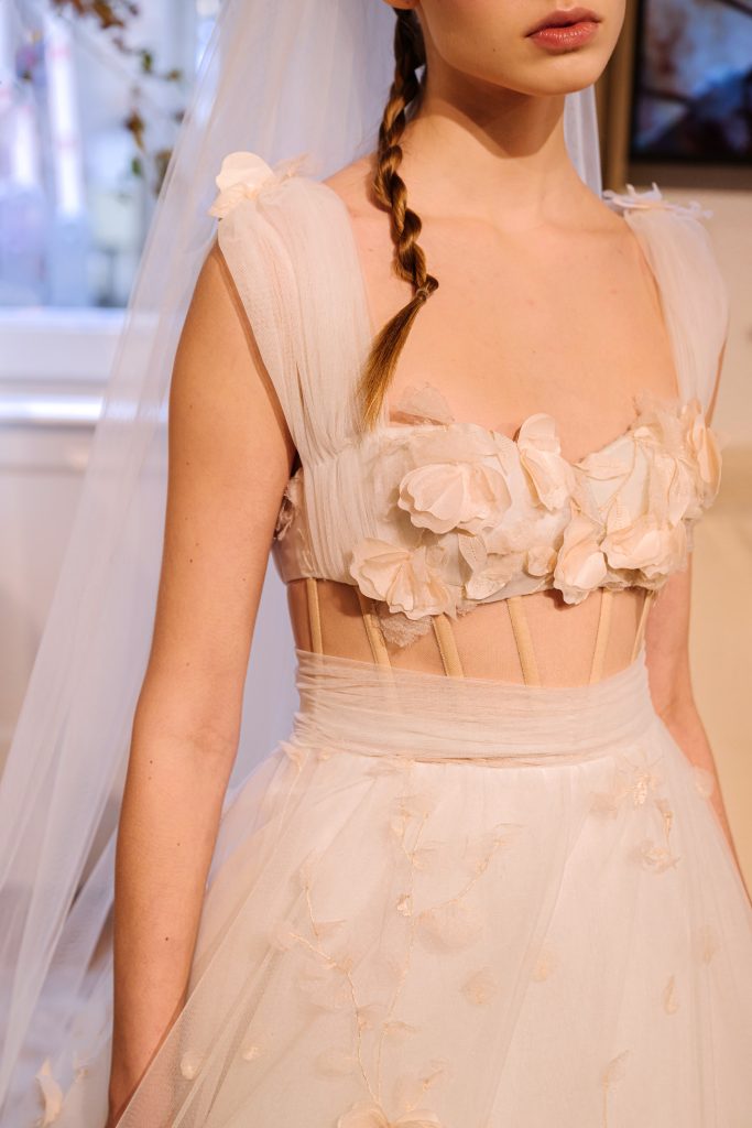 In questa foto, la modella indossa un abito romantico della collezione abiti da sposa Atelier Emé 2026, con un bustino semitrasparente decorato da fiori applicati in tessuto tono su tono. Le spalline morbide in tulle e la treccia lunga della modella completano il look etereo e femminile, perfetto per una sposa che sogna un’eleganza delicata.