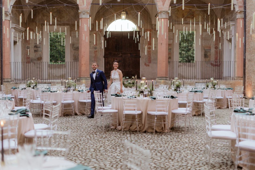 In questa foto dei Matrimoni Vincenzo Pioggia 2024 2025, gli sposi percorrono mano nella mano la navata centrale di una location allestita per il ricevimento: tavoli tondi, sedie trasparenti e candele sospese nell’aria creano un’atmosfera da sogno in un antico edificio con archi e colonne.