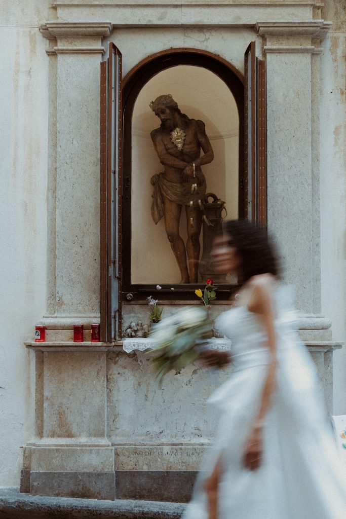 In questa foto, una sposa sfocata passa di fronte a un’edicola sacra con la statua di Cristo flagellato. Il contrasto tra il movimento moderno della sposa e la staticità della figura religiosa crea una potente narrazione visiva. Lo scatto rientra nella selezione Matrimoni Vincenzo Pioggia 2024 2025, dove ogni dettaglio racconta un’emozione.