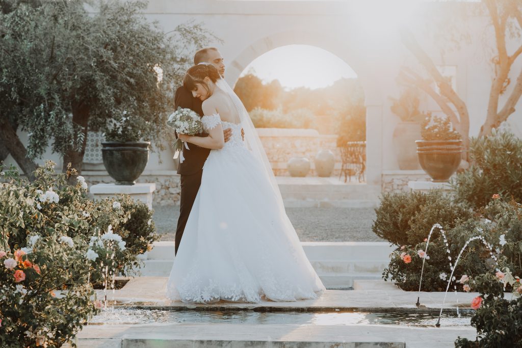 In questa foto dei Matrimoni Vincenzo Pioggia 2024 2025, gli sposi si abbracciano teneramente accanto a una fontana, circondati da fiori e piante in un’elegante corte mediterranea. Il sole al tramonto filtra dolcemente tra gli alberi, avvolgendo la scena in una luce calda e romantica.