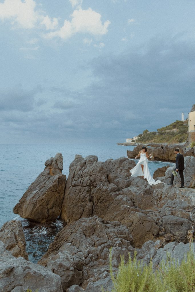 In questa foto, una sposa cammina tra gli scogli affacciati sul mare, seguita dallo sposo che la osserva con dolcezza. Il cielo è coperto da nuvole drammatiche, mentre l’acqua azzurra contrasta con la roccia grigia. Lo scatto fa parte della raccolta dei Matrimoni Vincenzo Pioggia 2024 2025, dove il paesaggio naturale diventa scenografia d’autore.