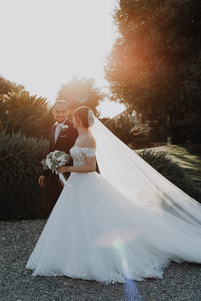 In questa foto dei Matrimoni Vincenzo Pioggia 2024 2025, gli sposi camminano sorridendo tra gli alberi, immersi in un parco baciato dalla luce del sole. L'abito della sposa, ampio e ricamato, si apre come una nuvola mentre il lungo velo si muove leggero al vento, trasmettendo un senso di gioia e complicità.