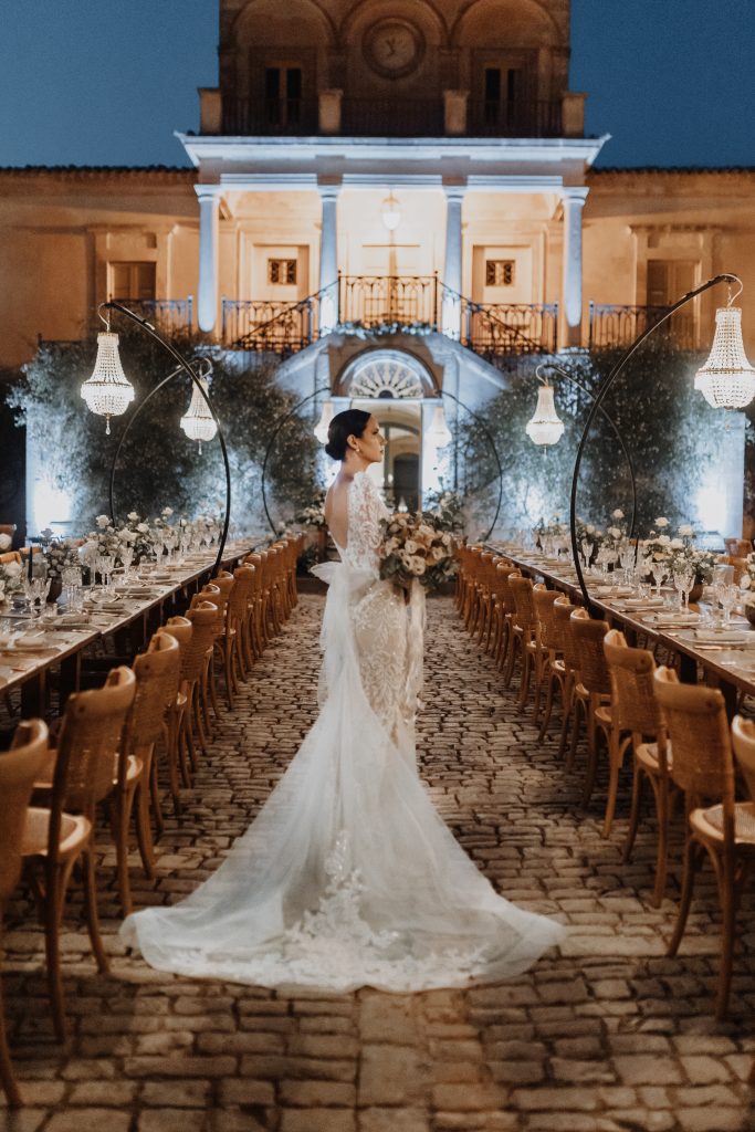 In questa foto, la sposa percorre un viale di pietra incorniciato da lunghe tavolate apparecchiate per il ricevimento serale. Sullo sfondo, una villa illuminata crea un’atmosfera scenografica, mentre lampadari sospesi aggiungono un tocco di magia. L’immagine è tratta dalla raccolta Matrimoni Vincenzo Pioggia 2024 2025.