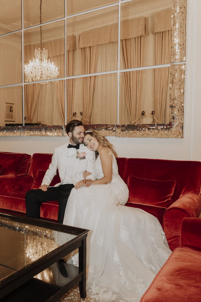 In questa foto, una coppia di sposi è seduta su un elegante divano rosso, immersa in un momento di dolce intimità. Lei indossa un abito da sposa con strascico e pizzo floreale, mentre lui è in smoking bianco con papillon nero. Si guardano e sorridono, appoggiati l’uno all’altra, sotto una luce calda riflessa da uno specchio con cornice dorata. L’immagine fa parte della raccolta Matrimoni Vincenzo Pioggia 2024 2025.