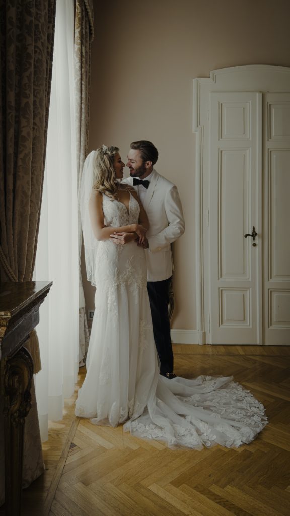 In questa foto, la sposa e lo sposo si abbracciano teneramente vicino a una finestra con tendaggi eleganti. L’abito da sposa presenta un profondo scollo a V e un lungo strascico decorato con ricami floreali. Lui, in giacca bianca e papillon nero, la guarda sorridendo. La luce naturale avvolge la scena in un’atmosfera intima e raffinata. Scatto della serie Matrimoni Vincenzo Pioggia 2024 2025.