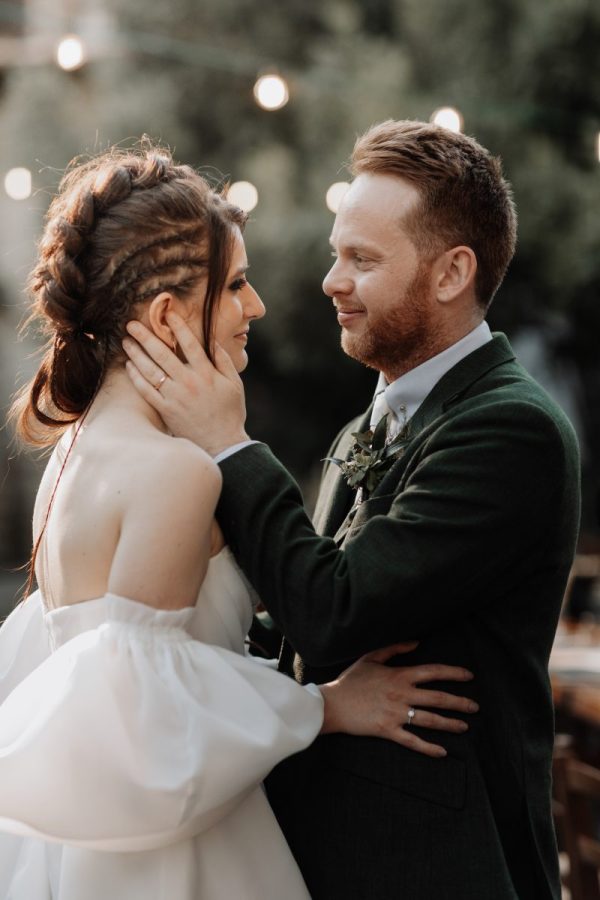In questa foto, un momento di intimità tra gli sposi: lui tiene il viso di lei tra le mani, guardandola con dolcezza. La sposa indossa un abito bianco con maniche vaporose, mentre lo sposo ha un completo verde scuro. La luce calda e le lampadine appese sullo sfondo creano un’atmosfera romantica e raccolta. La scena fa parte del progetto Matrimoni Vincenzo Pioggia 2024 2025.