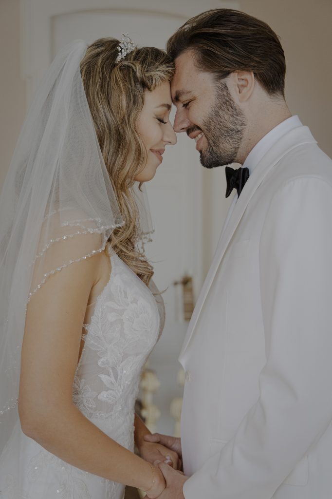 In questa foto, un primo piano dolce e romantico mostra gli sposi mentre si toccano la fronte con tenerezza, occhi chiusi e sorrisi sereni. La sposa indossa un velo con piccoli dettagli brillanti, e lo sposo è in giacca bianca. Si tengono per mano, trasmettendo complicità ed emozione. Un'immagine della collezione Matrimoni Vincenzo Pioggia 2024 2025.