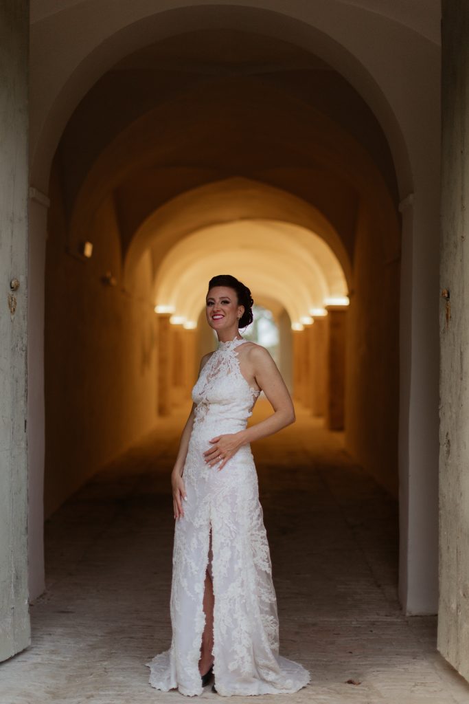 In questa foto dei Matrimoni Vincenzo Pioggia 2024 2025, la sposa è al centro di un elegante corridoio ad arcate illuminate, indossa un abito in pizzo con scollo all’americana e spacco centrale. Il suo sorriso illumina la scena, mentre la luce calda esalta la sua figura.