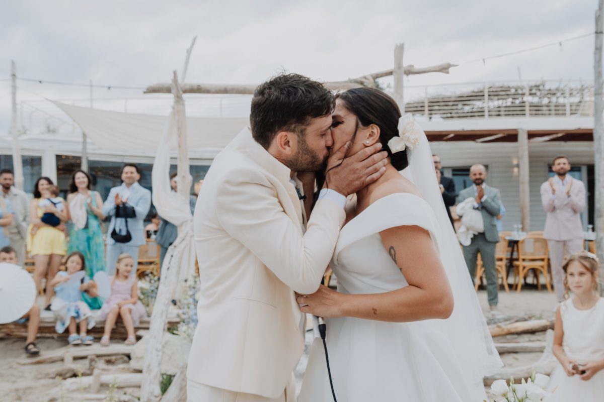 In questa foto, tratta dalla raccolta Matrimoni Vincenzo Pioggia 2024 2025, gli sposi si scambiano un bacio appassionato durante la cerimonia sulla spiaggia. Attorno a loro, amici e familiari applaudono con emozione, mentre sul tavolo dell’altare spicca una composizione floreale dai toni delicati. L’atmosfera è spontanea, autentica e carica di gioia condivisa.
