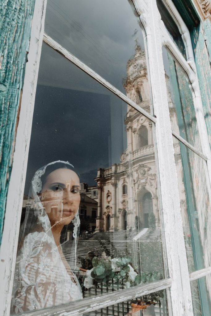 In questa foto, il riflesso di una sposa appare su una finestra antica, con il volto sereno incorniciato dal velo. Sul vetro si riflette anche una maestosa chiesa barocca, creando un gioco visivo tra interni ed esterni. Lo scatto fa parte della serie Matrimoni Vincenzo Pioggia 2024 2025.