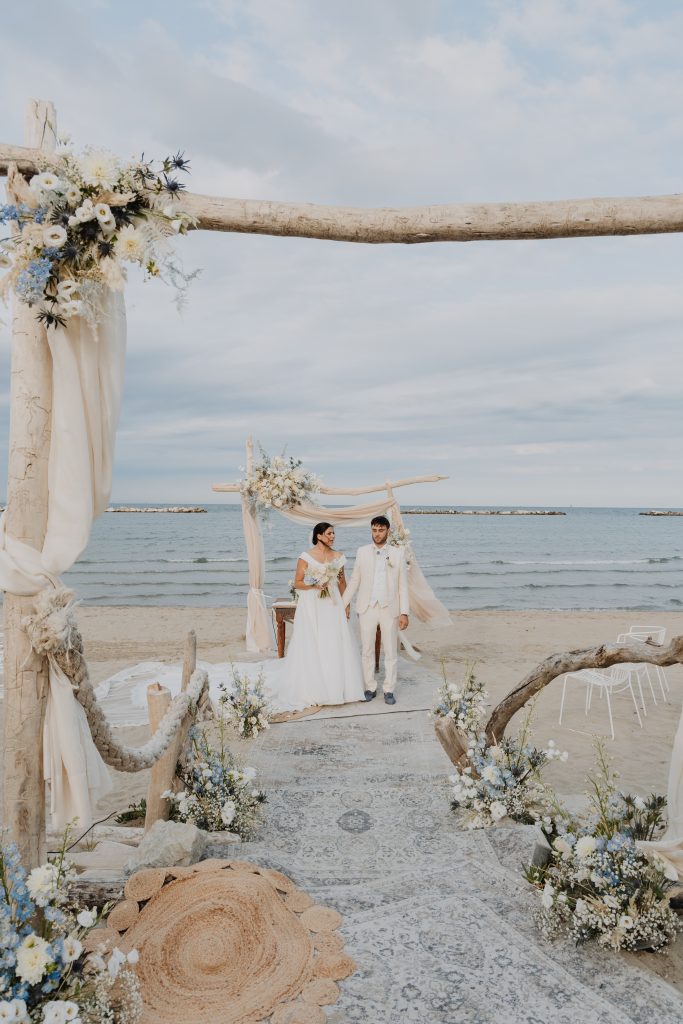 In questa foto dei Matrimoni Vincenzo Pioggia 2024 2025, gli sposi camminano mano nella mano sotto un arco decorato con fiori bianchi e blu, affacciati sul mare. L’ambientazione naturale e la palette cromatica tenue creano una cornice romantica e raffinata, perfettamente in linea con lo stile elegante del fotografo.
