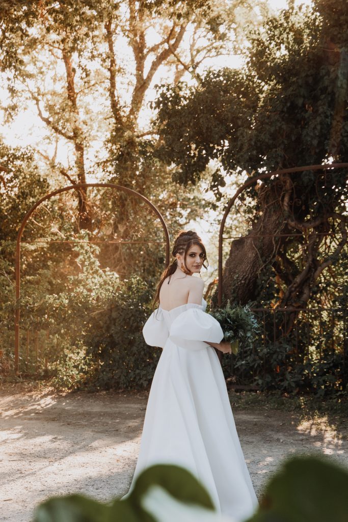 In questa foto, la sposa cammina tra alberi e archi di ferro, voltandosi verso l’obiettivo con uno sguardo elegante e intenso. Indossa un abito bianco semplice ma scenografico e tiene in mano un bouquet di foglie verdi. La luce dorata filtra tra gli alberi, esaltando l’atmosfera sognante. Uno scatto del progetto Matrimoni Vincenzo Pioggia 2024 2025.