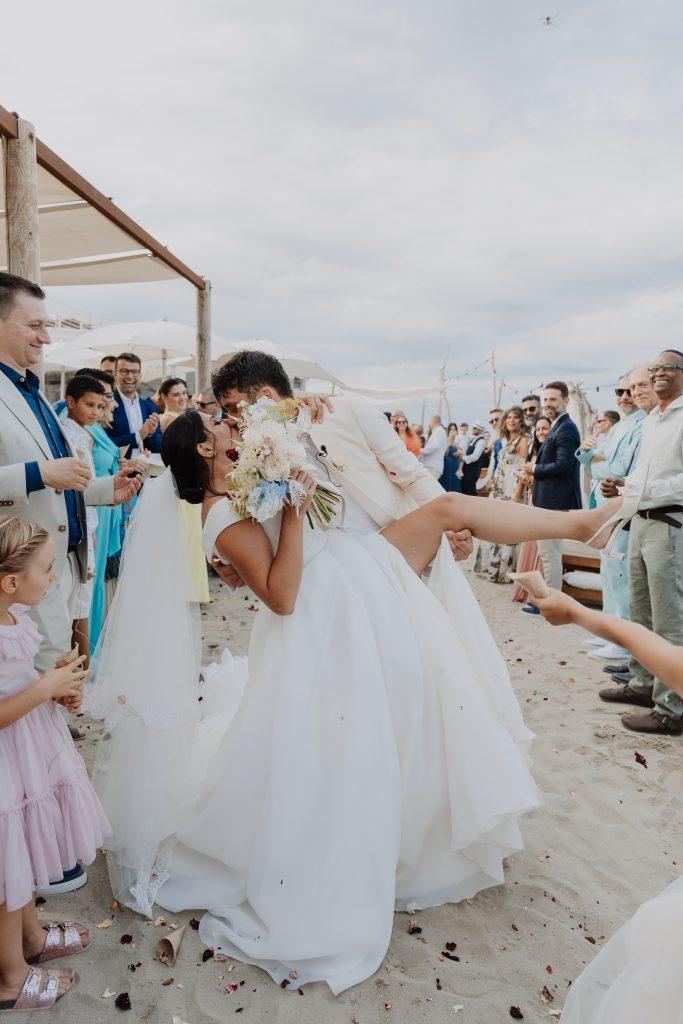 In questa foto realizzata per i Matrimoni Vincenzo Pioggia 2024 2025, lo sposo solleva la sposa con un gesto giocoso e romantico subito dopo la cerimonia. Gli invitati li circondano sorridendo, creando un momento di festa collettiva sulla sabbia, tra petali lanciati e risate. L’immagine cattura un’esplosione di felicità autentica.