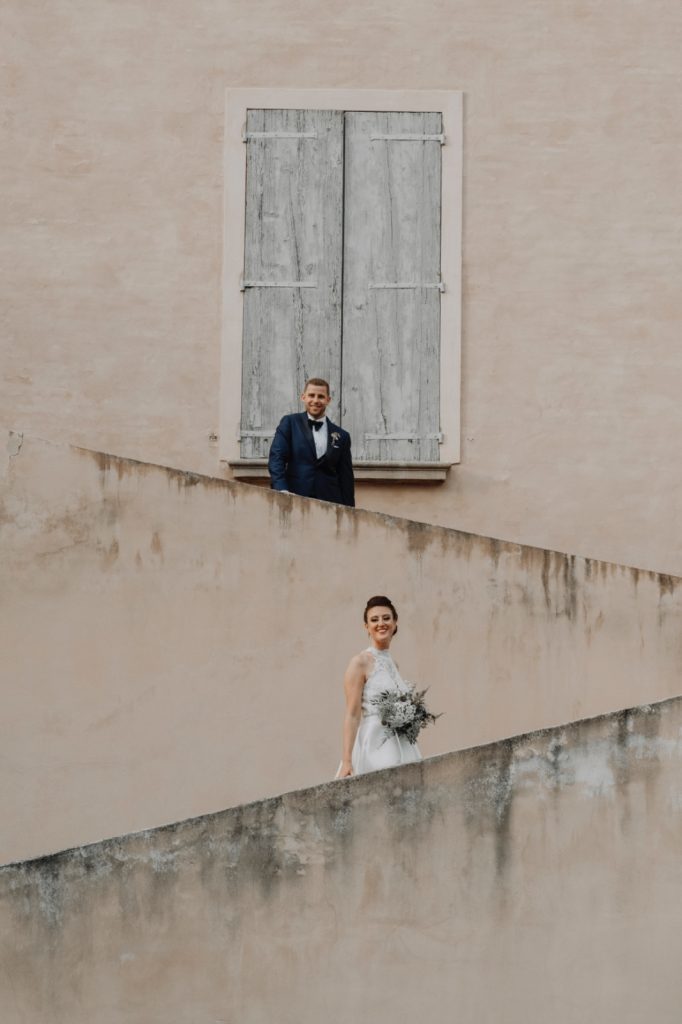 In questa foto dei Matrimoni Vincenzo Pioggia 2024 2025, la sposa e lo sposo sono immortalati su due livelli diversi di una scalinata dai toni neutri: lui in alto accanto a una finestra chiusa in legno, lei più in basso, sorridente e radiosa con il bouquet tra le mani. Un’inquadratura pulita e geometrica che gioca con le linee architettoniche.