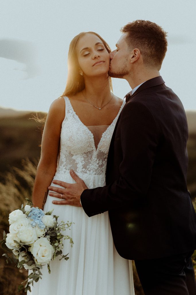 In questa foto, una sposa con un abito in pizzo bianco viene teneramente baciata sulla guancia dal suo compagno, vestito con un abito scuro. Lei tiene tra le mani un bouquet di rose bianche e azzurre, mentre il tramonto dorato alle loro spalle crea un'atmosfera calda e romantica. Lo stile è intimo e naturale, tipico degli scatti dei Matrimoni Vincenzo Pioggia 2024 2025.
