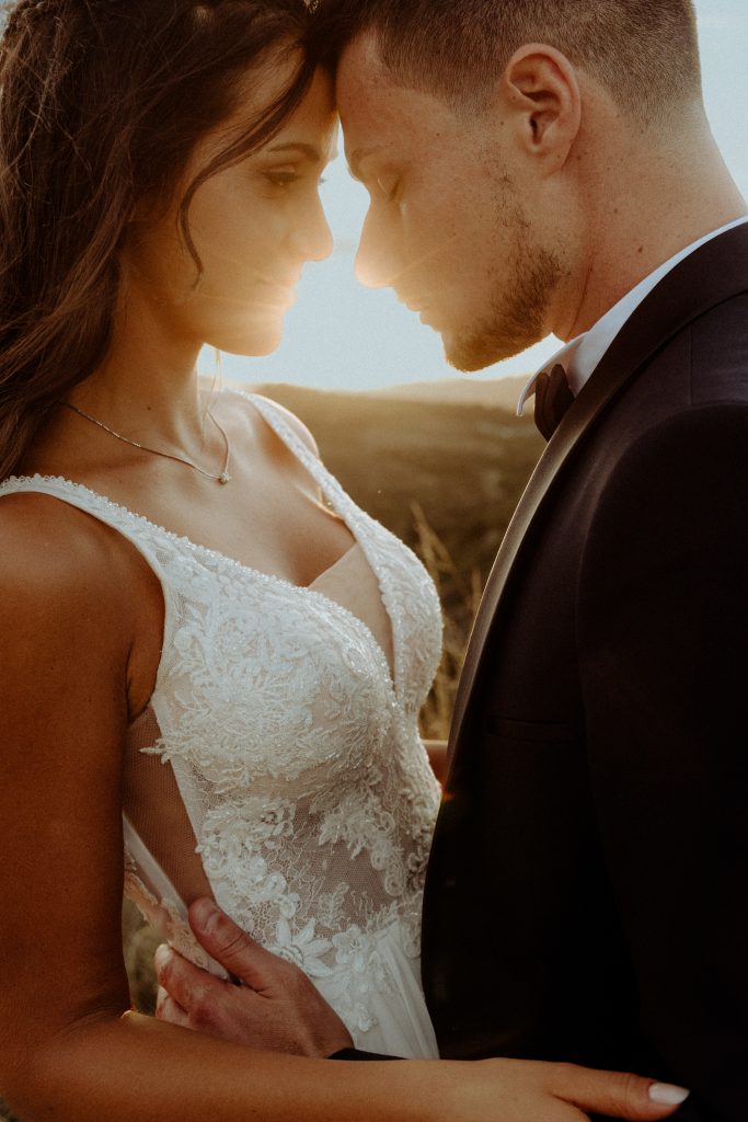 In questa foto, gli sposi si guardano intensamente negli occhi, con le fronti che si toccano. La luce del sole filtra tra i loro volti, creando un’atmosfera carica di emozione. Il dettaglio sul corpetto in pizzo dell’abito da sposa mette in risalto l’eleganza sartoriale, in perfetto stile Matrimoni Vincenzo Pioggia 2024 2025.