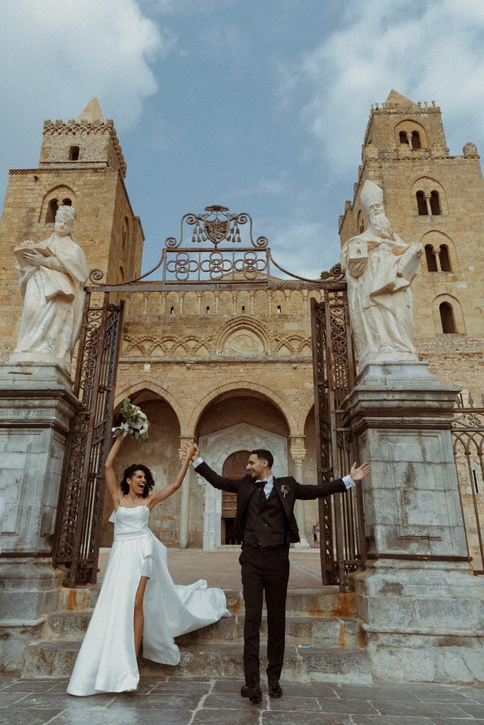 In questa foto, una coppia di sposi esulta sotto il maestoso arco d’ingresso della Cattedrale di Cefalù, con le braccia alzate in un gesto di gioia. Le statue e le torri antiche incorniciano il momento, esaltando l’armonia tra storia e amore. L’immagine è tratta dai Matrimoni Vincenzo Pioggia 2024 2025.