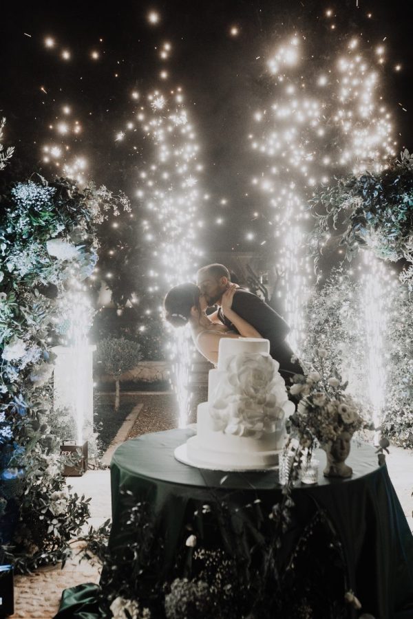 In questa foto dei Matrimoni Vincenzo Pioggia 2024 2025, una coppia di sposi si bacia davanti alla torta nuziale, circondata da un'esplosione di scintille e fuochi d'artificio scenografici. Il momento, immortalato di sera, è reso ancora più magico dalla luce calda e dai giochi luminosi che incorniciano gli sposi, creando un’atmosfera da fiaba.