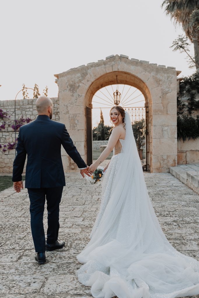 In questa foto, gli sposi camminano mano nella mano verso un grande portale in pietra, con la sposa che si volta e sorride felice verso la fotocamera. Il tramonto illumina la scena con una luce dorata e delicata. Lo scatto immortala la gioia e la complicità tipica dei Matrimoni Vincenzo Pioggia 2024 2025.