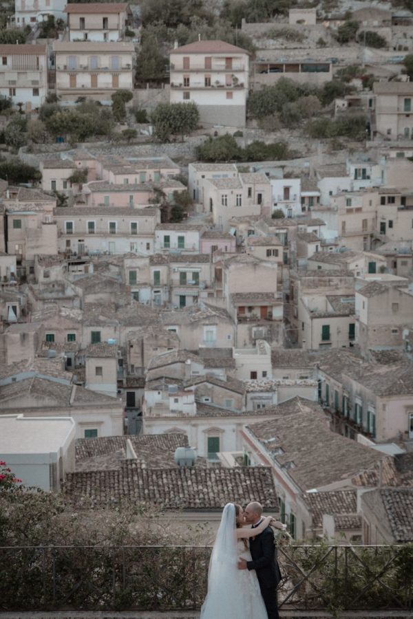 In questa foto, gli sposi si abbracciano e si baciano dolcemente con vista panoramica su un borgo antico, fatto di tetti in pietra e case color pastello. Il paesaggio urbano, sfocato sullo sfondo, valorizza l’intimità della coppia. L’immagine è uno degli scatti iconici dei Matrimoni Vincenzo Pioggia 2024 2025.