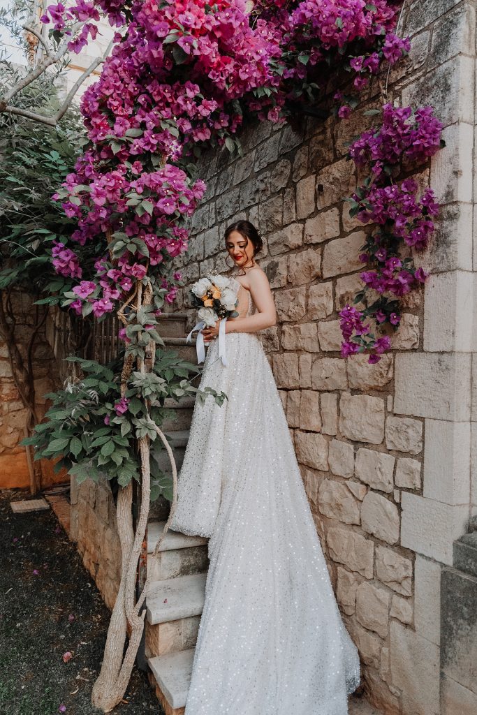 In questa foto, una sposa in abito scintillante si trova su una scalinata in pietra, avvolta da un arco naturale di bouganville fucsia. Tiene in mano un bouquet dai toni delicati e sorride dolcemente, mentre la luce calda del tramonto esalta i dettagli dell'abito. Uno scatto romantico che rappresenta perfettamente l’eleganza dei Matrimoni Vincenzo Pioggia 2024 2025.