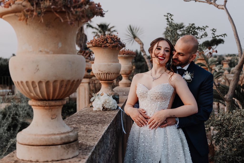 In questa foto, la sposa e lo sposo sono abbracciati su una terrazza ornata da grandi vasi in pietra. Lei, raggiante e sorridente, indossa un abito bianco tempestato di perle e guarda l’obiettivo; lui la stringe da dietro con sguardo innamorato. Un momento di intimità autentica nei Matrimoni Vincenzo Pioggia 2024 2025.