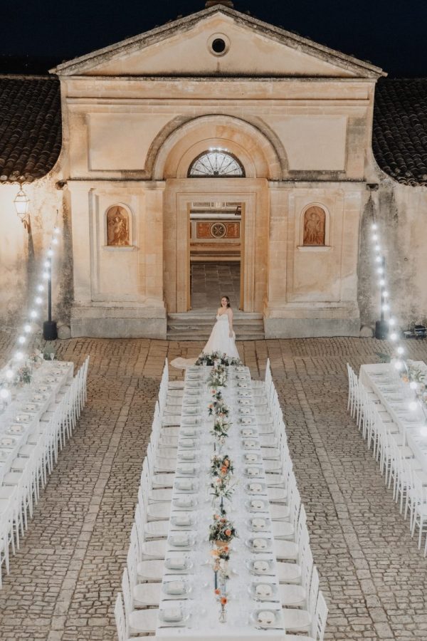 In questa foto, una sposa in abito bianco posa al centro di un suggestivo allestimento per la cena di nozze, incorniciato da una corte in pietra con tavoli lunghi e decorazioni floreali. Le luci calde e la facciata di un’antica chiesa sullo sfondo creano un’atmosfera romantica e senza tempo. L’immagine fa parte della selezione dei Matrimoni Vincenzo Pioggia 2024 2025.