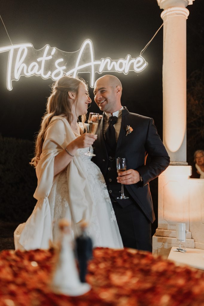 In questa foto, una coppia di sposi brinda sorridente davanti a un’insegna luminosa che recita “That’s Amore”. Lui in completo scuro, lei in abito bianco con mantella sulle spalle: i due si guardano con complicità durante il ricevimento serale. Questa scena emozionante è uno dei momenti speciali dei Matrimoni Vincenzo Pioggia 2024 2025.