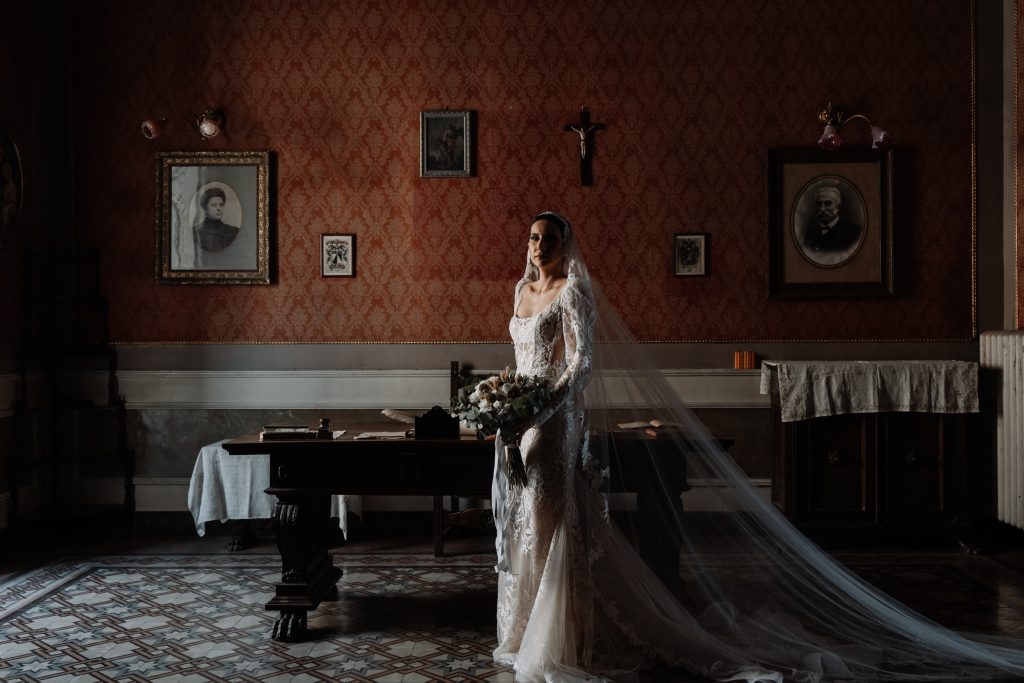 In questa foto, una sposa in abito di pizzo e lungo velo sosta in una stanza antica dai toni caldi, con pareti damascate rosse e pavimento decorato. La luce entra da una grande finestra laterale, accarezzando la figura centrale. È un'immagine elegante e silenziosa, che fa parte del progetto fotografico Matrimoni Vincenzo Pioggia 2024 2025.