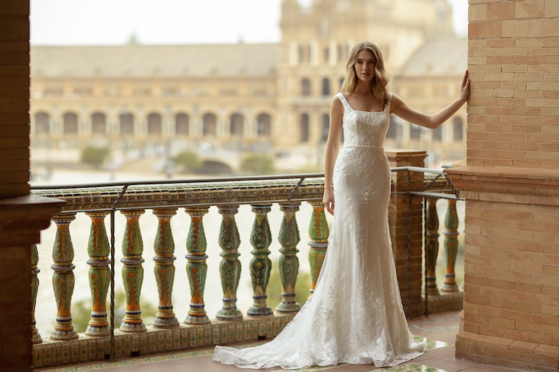 In questa foto, una modella posa indossando un abito da sposa di Monica Loretti con una linea scivolata, scollo quadrato e dettagli in pizzo