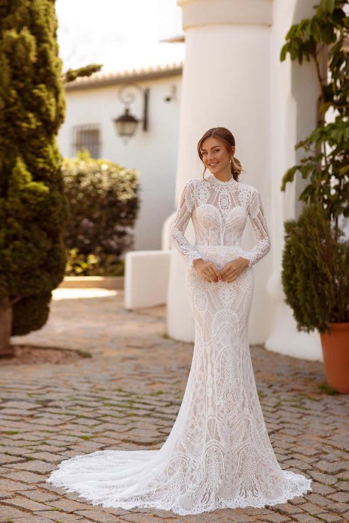 In questa foto, una modella posa indossando un abito a semi-sirena di Monica Loretti in pizzo macramè con maniche e scollatura girocollo