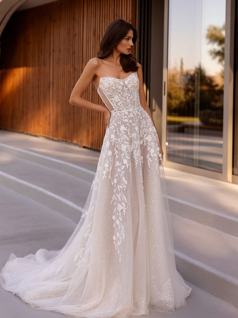 In questa foto, una modella indossa un raffinato abito da sposa con taglio scivolato, perfetto esempio della tendenza abiti da sposa scivolati 2026. Il vestito, senza spalline, è impreziosito da ricami floreali delicati e luminosi su un tessuto semi-trasparente che crea un effetto etereo e romantico. La gonna ampia ma fluida cade con naturalezza, accompagnata da un lungo strascico, mentre il corpetto sagomato mette in risalto la figura della sposa. Sullo sfondo, un’elegante architettura contemporanea con pareti in legno e grandi vetrate.