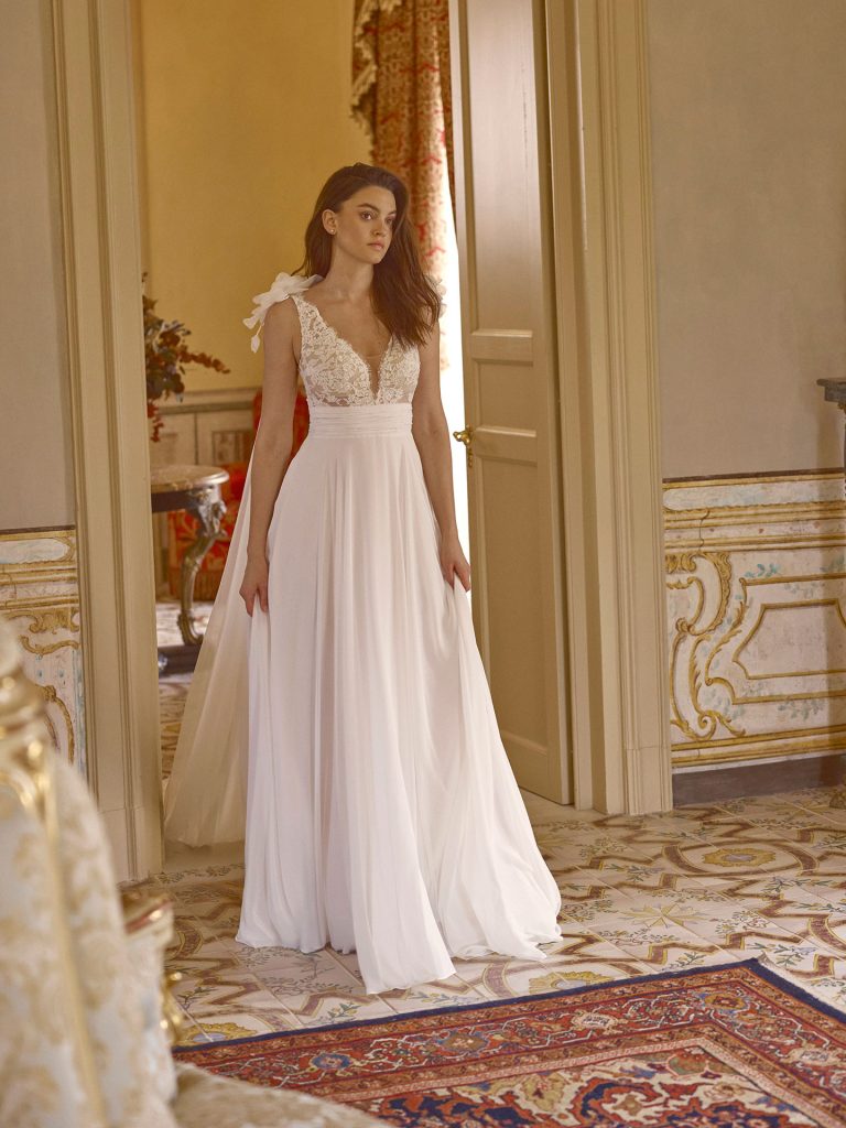 In questa foto, una sposa cammina in un ambiente riccamente decorato, indossando un abito con corpetto in pizzo e spalline a fiocco. La gonna leggera e scivolata esalta la silhouette con naturalezza, incarnando l’essenza romantica degli abiti da sposa scivolati 2026.