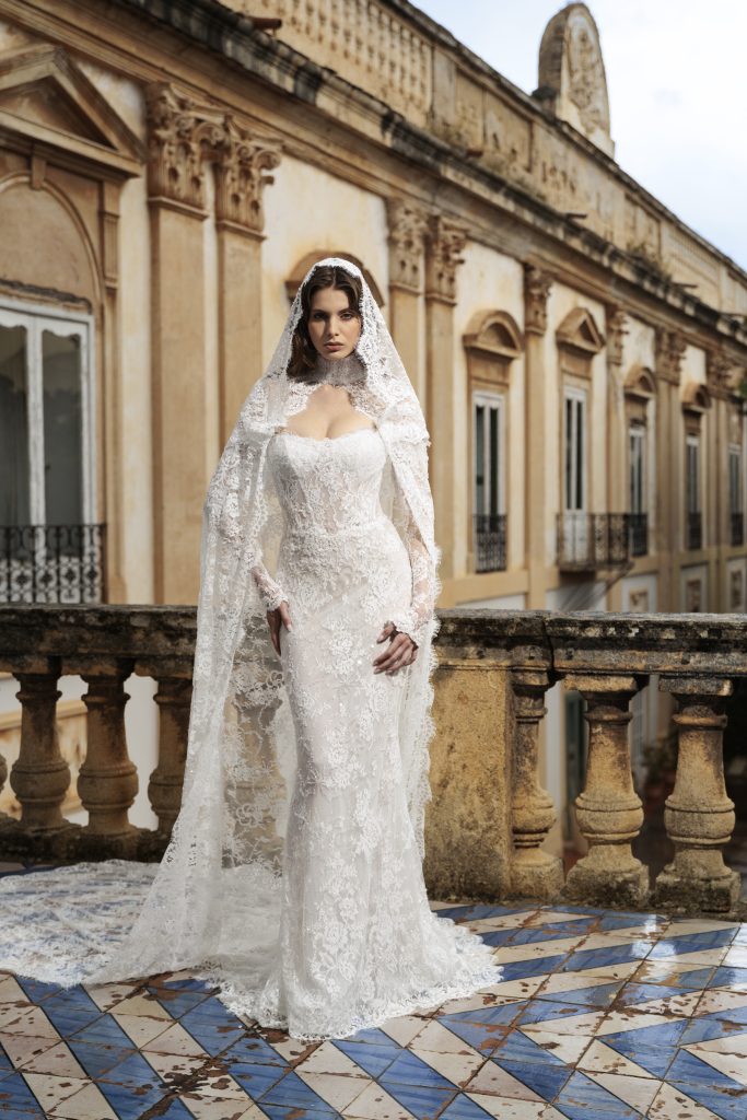 In questa foto, una sposa si trova su una terrazza in pietra, con un abito in pizzo interamente ricamato e un velo coordinato con cappuccio in stile antico. La linea affusolata dell’abito abbraccia il corpo con eleganza, in perfetto stile abiti da sposa scivolati.