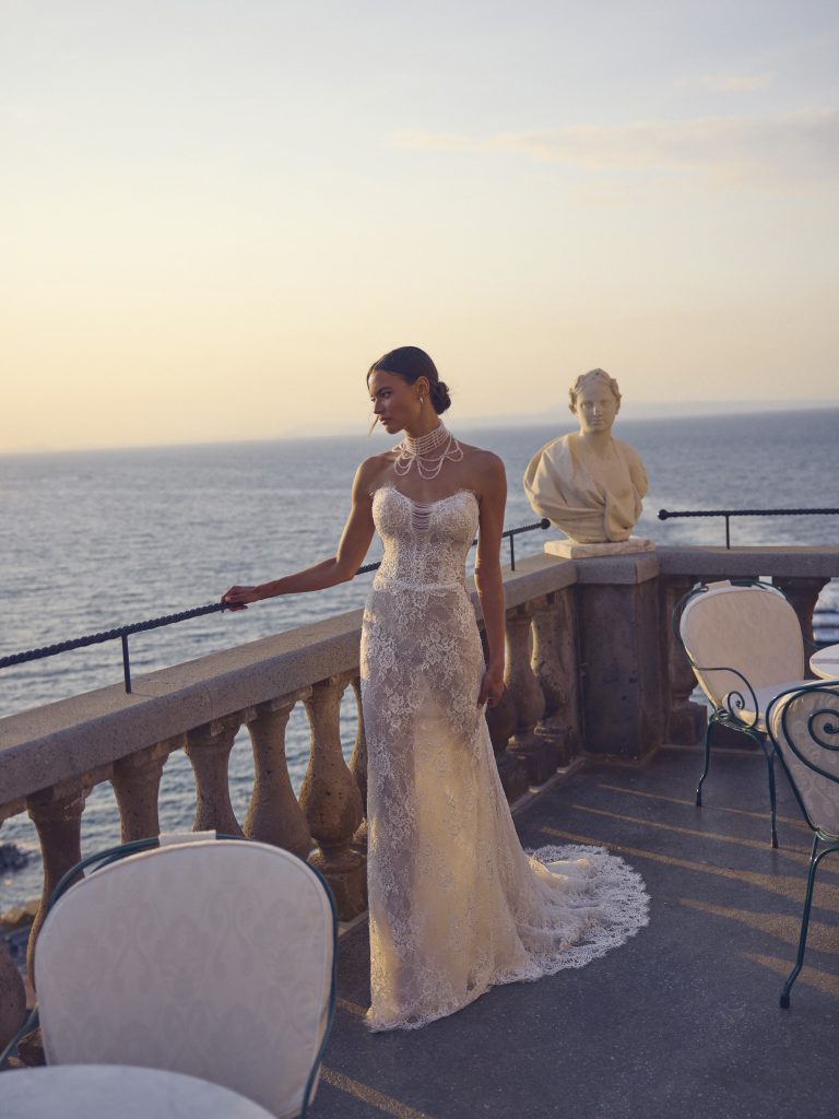 In questa foto, una sposa si trova su una terrazza con vista mare al tramonto, indossando un abito a sirena in pizzo trasparente e un elaborato collier a più fili. La silhouette morbida e sensuale delinea perfettamente lo stile degli abiti da sposa scivolati più glamour.