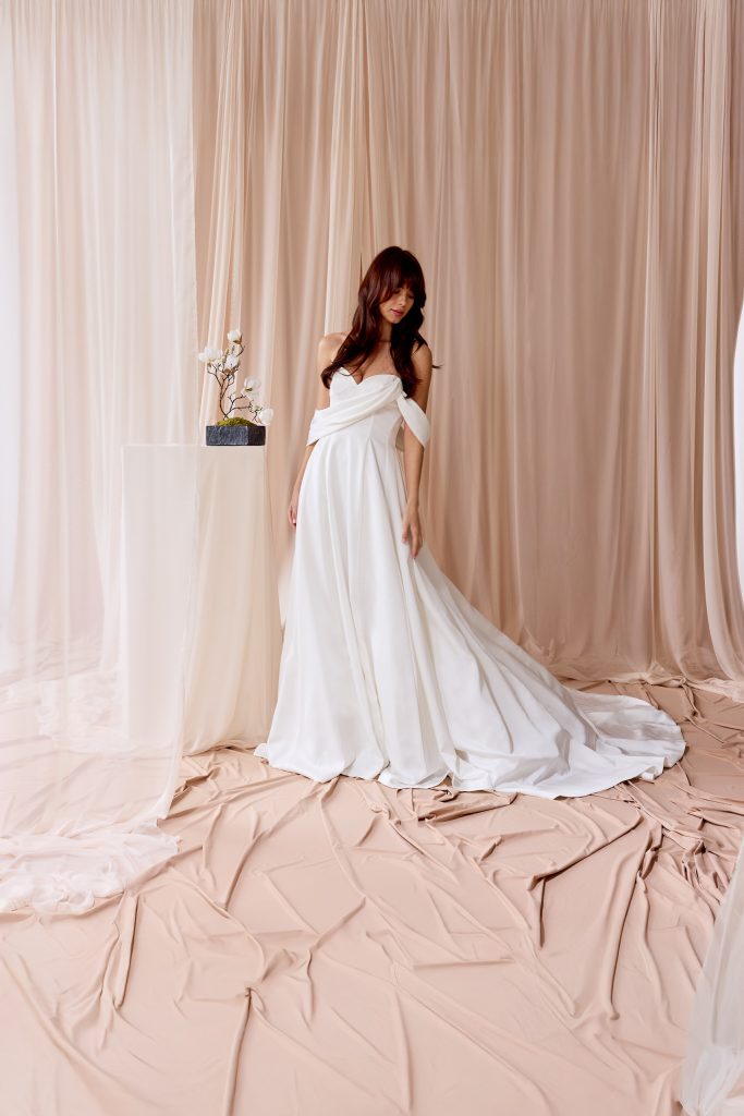 In questa foto, una modella indossa un elegante abito da sposa scivolato 2026 caratterizzato da un corpetto drappeggiato e spalle scoperte. La scena si svolge in un ambiente raffinato dai toni neutri, con tendaggi color cipria e un piccolo tavolo decorato con fiori bianchi. Il tessuto dell’abito cade fluido fino a terra, creando una silhouette morbida e romantica.