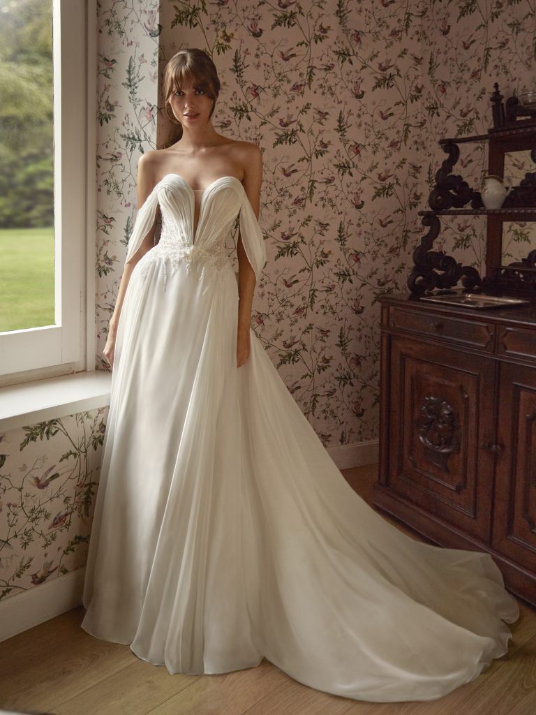 In questa foto, una sposa sosta accanto a una finestra in una stanza con carta da parati floreale, indossando un abito con corpetto drappeggiato, spalle scoperte e gonna fluente in chiffon. Il look etereo e leggero è perfetto per chi cerca abiti da sposa scivolati dallo stile boho chic.
