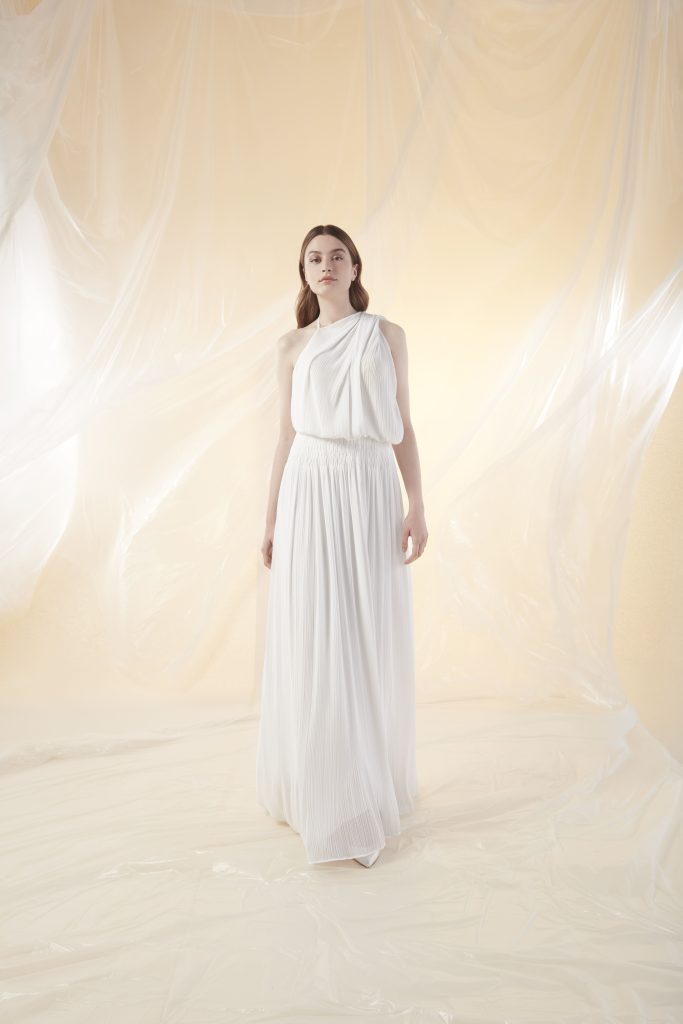 In questa foto, una modella posa su uno sfondo chiaro e neutro, con indosso un abito monospalla con top drappeggiato e gonna morbida in tessuto plissé. Il design moderno e raffinato rappresenta una delle interpretazioni più essenziali degli abiti da sposa scivolati 2026.