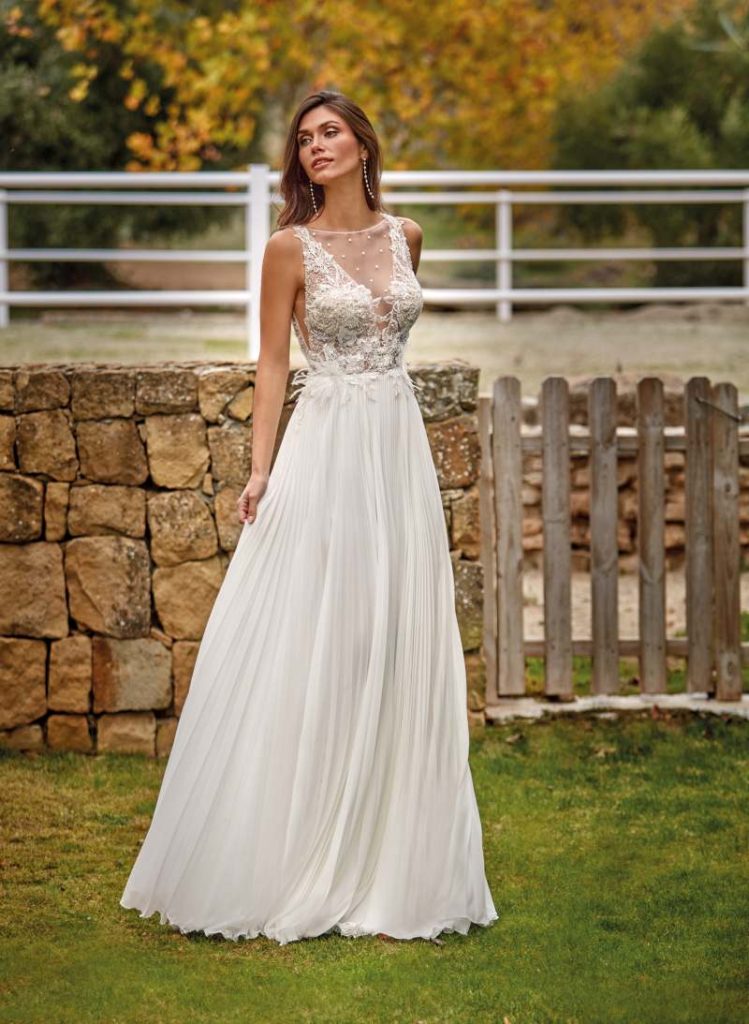 In questa foto, una modella indossa uno degli abiti da sposa scivolati 2026, perfetto per chi sogna un look etereo e romantico. Il vestito, caratterizzato da un corpino ricamato con dettagli in pizzo e una gonna in chiffon leggero e plissettato, si muove con grazia sul prato verde, evocando un’eleganza naturale e raffinata. Lo scollo illusione e le trasparenze delicate conferiscono un tocco di sensualità discreta.